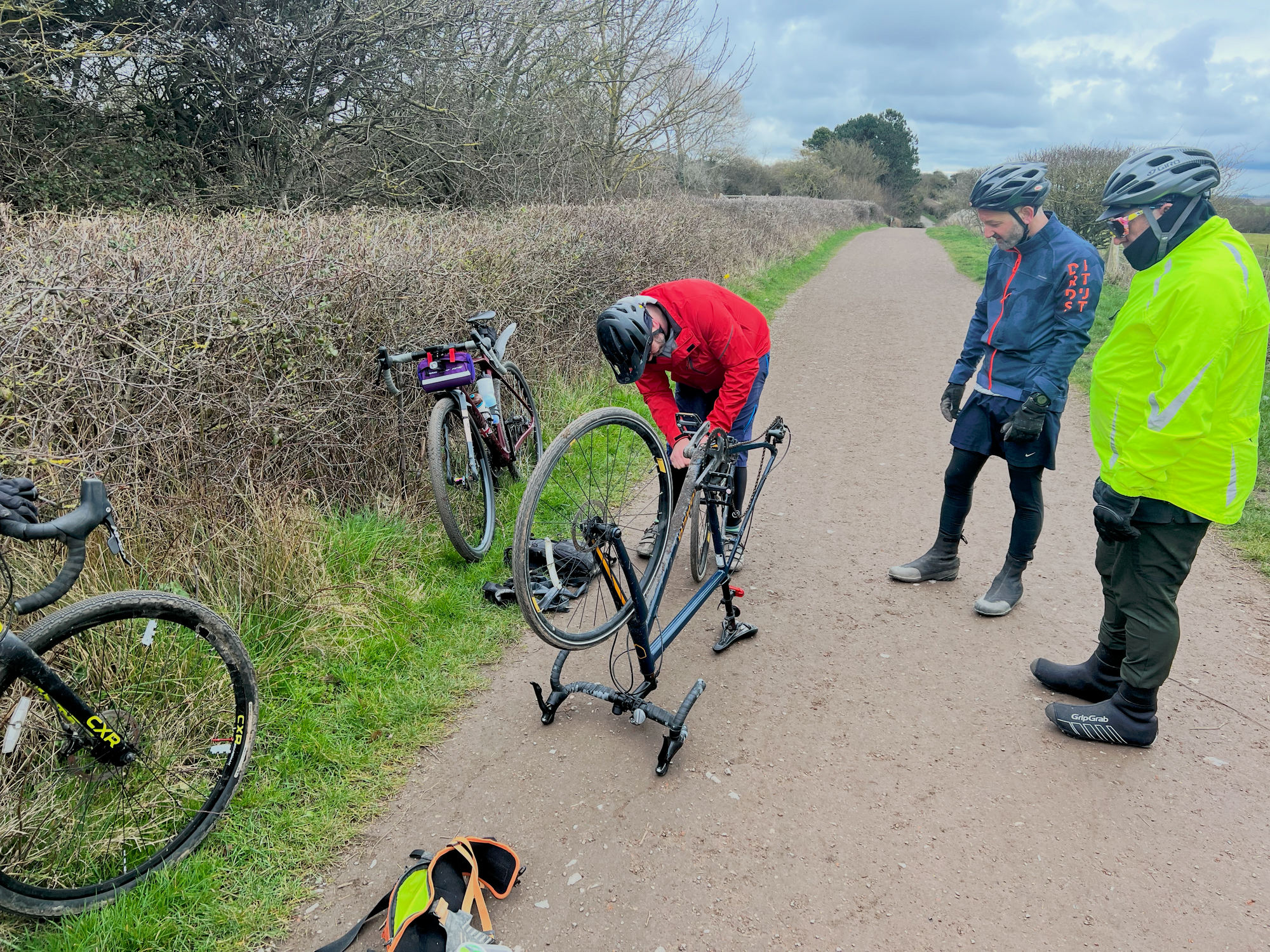 2 men in cycling cothing watch a man fixing a puncture on his bike.