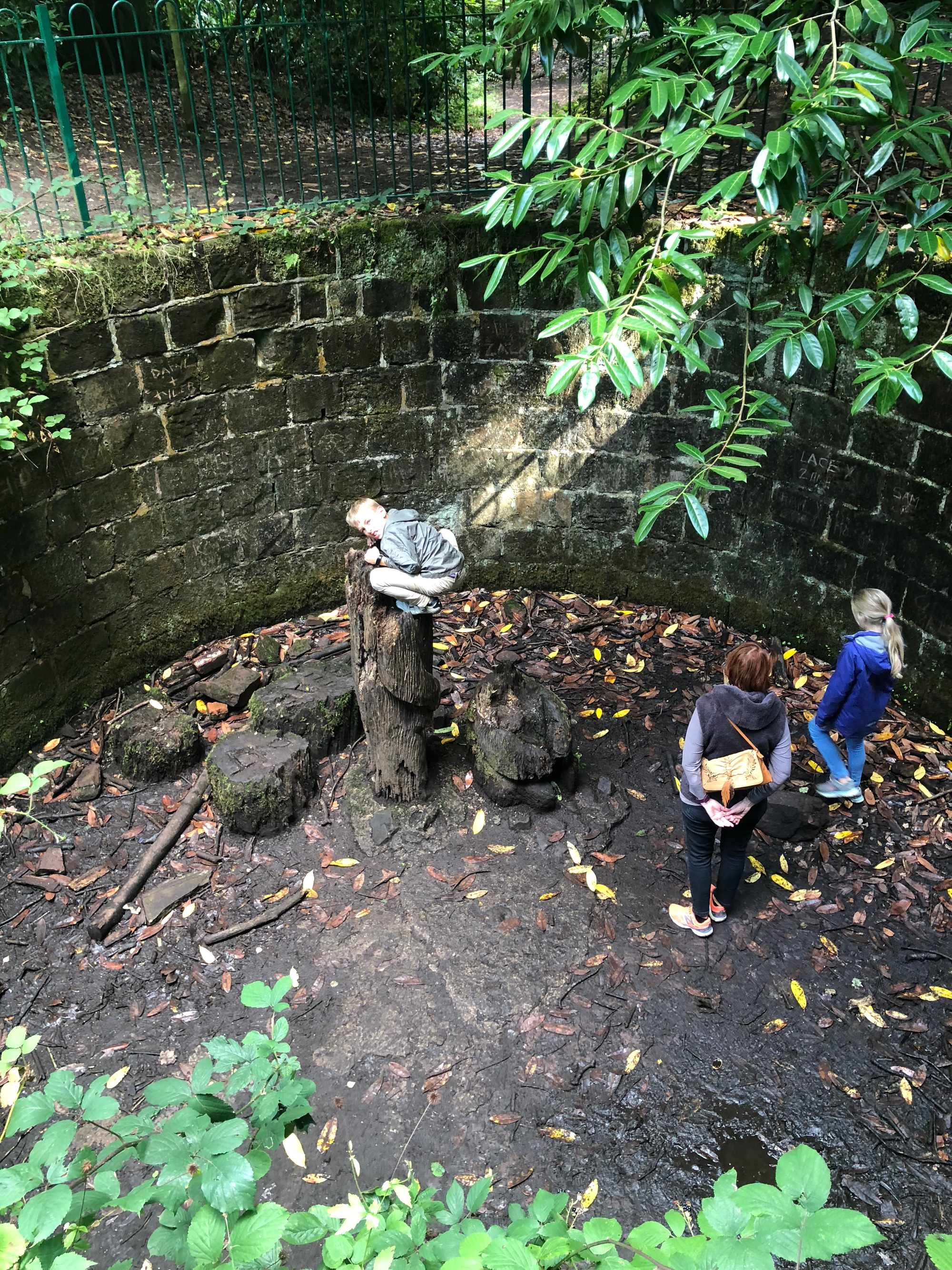 2 children and an adult in a pit.  There are thick sandstone walls towering above and a green fence around the top.  The photographer is looking down on the scene.