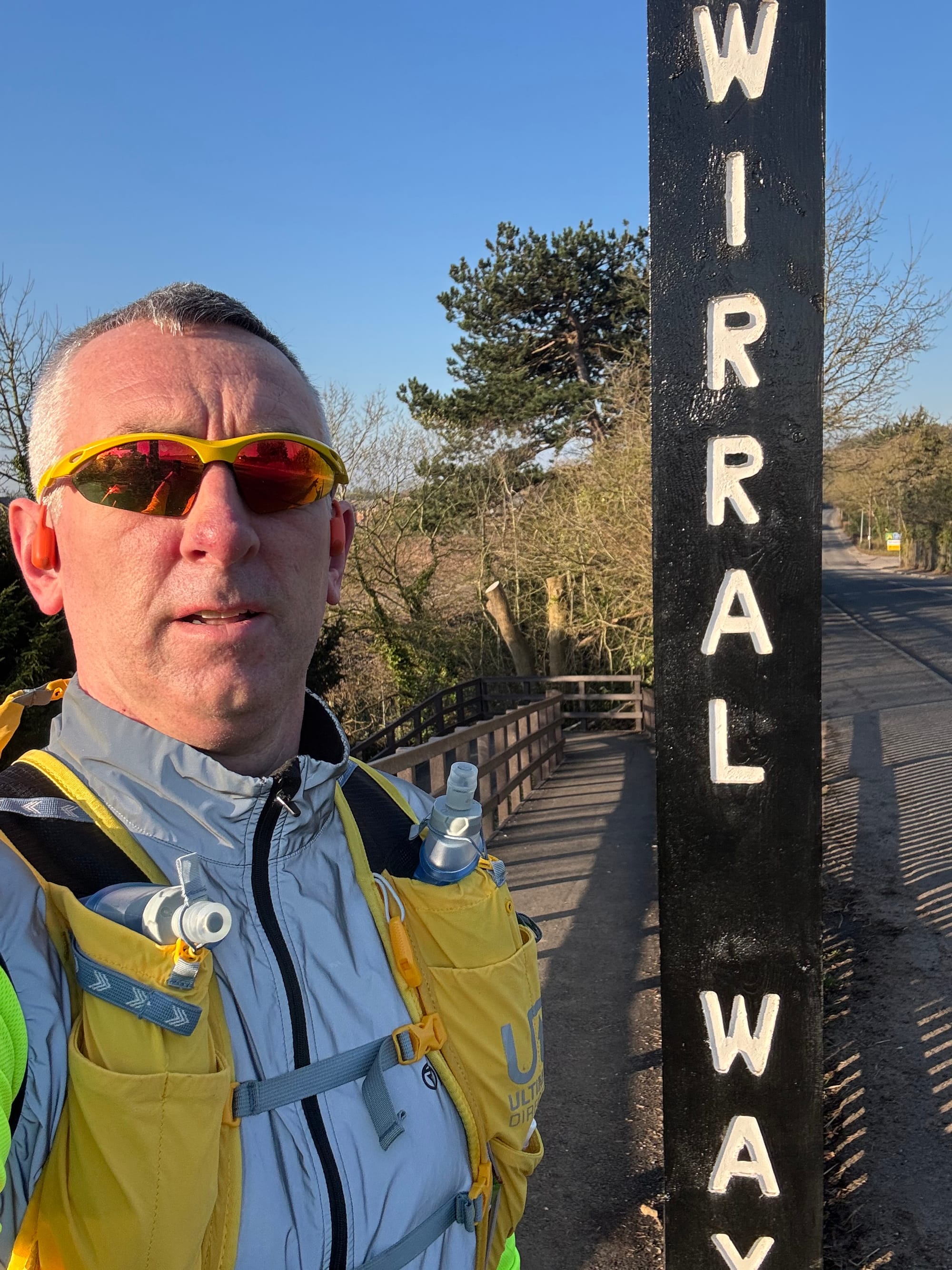 Man standing next to a sign saying The Wirral Way.  He is in a grey jacket, yellow running vest and wears shades.
