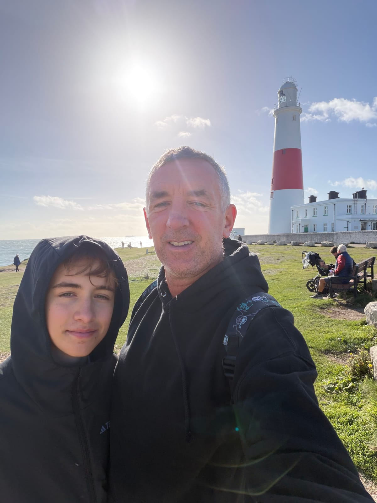 Man and Boy standing in front of Portland Bill lighthouse. The lighthouse’s white with a red strip around the middle. The sun is shining